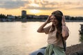 Young hipster millennial woman sitting on riverside looking at city listening music on wireless headphones enjoying moment, life Royalty Free Stock Photo