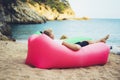 Young hipster relaxing on coastline beach on inflatable lazy air pouffe sofa, person tourist enjoy sunny day on background coast