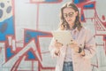 Young hipster woman in glasses stands on city street and uses tablet computer. In background wall with graffiti. Royalty Free Stock Photo
