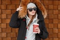 Young hipster woman in a knitted gray hat in a stylish winter jacket with fur in a warm blue sweatshirt holding a cup of coffee Royalty Free Stock Photo