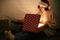 Young hipster woman in santa hat opening magical present in dark room and smiling. Merry Christmas. Stylish happy girl in cozy Royalty Free Stock Photo