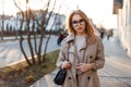 Young hipster woman in a stylish spring coat in fashionable glasses in a white T-shirt in a black leather handbag walking along