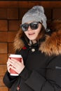 Young hipster woman in a warm knitted hat in sunglasses in a black stylish black jacket with a fur hood and a cup of hot coffee Royalty Free Stock Photo