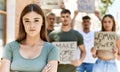 Young hispanic activist woman with arms crossed gesture standing with a group of protesters holding banner protesting at the city Royalty Free Stock Photo