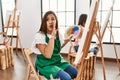 Young hispanic artist women painting on canvas at art studio hand on mouth telling secret rumor, whispering malicious talk Royalty Free Stock Photo