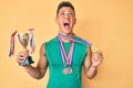 Young hispanic boy holding champion trophy wearing medals angry and mad screaming frustrated and furious, shouting with anger Royalty Free Stock Photo