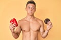 Young hispanic boy shirtless holding red pepper and donut puffing cheeks with funny face