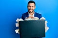 Young hispanic businessman holding briefcase full of dollars sticking tongue out happy with funny expression Royalty Free Stock Photo