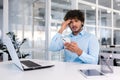 Young hispanic businessman sitting at desk in office, holding phone, worriedly holding head and looking at camera Royalty Free Stock Photo