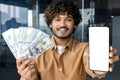 Young hispanic businessman at workplace smile and look at camera, man shows blank white phone screen and cash money Royalty Free Stock Photo