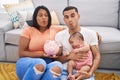 Young hispanic couple with baby holding piggy bank making fish face with mouth and squinting eyes, crazy and comical