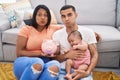 Young hispanic couple with baby holding piggy bank puffing cheeks with funny face