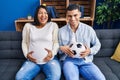 Young hispanic couple expecting a baby sitting on the sofa holding ball and tummy smiling and laughing hard out loud because funny Royalty Free Stock Photo