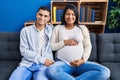 Young hispanic couple expecting a baby sitting on the sofa at home looking positive and happy standing and smiling with a Royalty Free Stock Photo