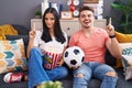 Young hispanic couple football hooligans holding ball and eating popcorn smiling with an idea or question pointing finger with