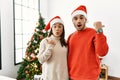 Young hispanic couple standing by christmas tree surprised pointing with hand finger to the side, open mouth amazed expression Royalty Free Stock Photo