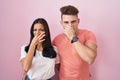 Young hispanic couple standing over pink background smelling something stinky and disgusting, intolerable smell, holding breath Royalty Free Stock Photo