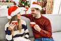 Young hispanic couple wearing christmas hat drinking coffee sitting on the sofa at home Royalty Free Stock Photo