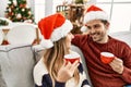 Young hispanic couple wearing christmas hat drinking coffee sitting on the sofa at home Royalty Free Stock Photo