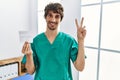 Young hispanic doctor man holding covid record card smiling looking to the camera showing fingers doing victory sign