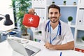 Young hispanic doctor man holding first aid kit looking positive and happy standing and smiling with a confident smile showing Royalty Free Stock Photo