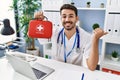 Young hispanic doctor man holding first aid kit pointing thumb up to the side smiling happy with open mouth Royalty Free Stock Photo