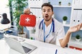 Young hispanic doctor man holding first aid kit scared and amazed with open mouth for surprise, disbelief face Royalty Free Stock Photo