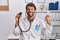 Young hispanic doctor man wearing doctor uniform holding stethoscope at clinic pointing thumb up to the side smiling happy with Royalty Free Stock Photo