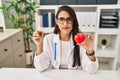 Young hispanic doctor woman holding heart and cbd oil relaxed with serious expression on face