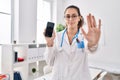 Young hispanic doctor woman holding smartphone showing screen with open hand doing stop sign with serious and confident Royalty Free Stock Photo