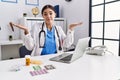 Young hispanic doctor woman wearing doctor uniform working at the clinic clueless and confused expression with arms and hands Royalty Free Stock Photo