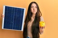 Young hispanic girl holding photovoltaic solar panel and piggy bank puffing cheeks with funny face