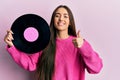 Young hispanic girl holding vinyl disc smiling happy and positive, thumb up doing excellent and approval sign Royalty Free Stock Photo