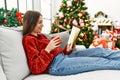 Young hispanic girl reading book and drinking coffee sitting on the sofa by christmas tree at home Royalty Free Stock Photo