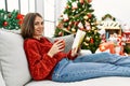 Young hispanic girl reading book and drinking coffee sitting on the sofa by christmas tree at home Royalty Free Stock Photo