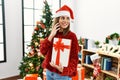 Young hispanic girl talking on the smartphone and holding gift standing by christmas tree at home Royalty Free Stock Photo