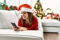 Young hispanic girl using smartphone lying on the sofa by christmas tree at home Royalty Free Stock Photo