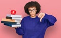 Young hispanic girl wearing glasses holding books and red apple with angry face, negative sign showing dislike with thumbs down, Royalty Free Stock Photo