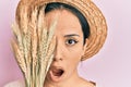 Young hispanic girl wearing summer hat holding spike wheat over eye afraid and shocked with surprise and amazed expression, fear Royalty Free Stock Photo