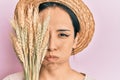 Young hispanic girl wearing summer hat holding spike wheat over eye puffing cheeks with funny face Royalty Free Stock Photo