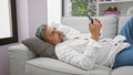 Young hispanic grey-haired man using smartphone lying on sofa at home Royalty Free Stock Photo
