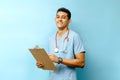 Young Hispanic male doctor holding a medical chart and a pen on a blue background. Royalty Free Stock Photo