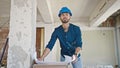 Young hispanic man architect wearing hardhat looking at blueprints at construction site