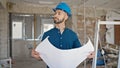 Young hispanic man architect wearing hardhat looking at blueprints at construction site