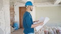 Young hispanic man architect wearing hardhat looking at blueprints at construction site