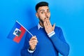 Young hispanic man with beard holding haiti flag covering mouth with hand, shocked and afraid for mistake