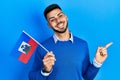 Young hispanic man with beard holding haiti flag smiling happy pointing with hand and finger to the side