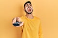 Young hispanic man with beard holding television remote control angry and mad screaming frustrated and furious, shouting with Royalty Free Stock Photo