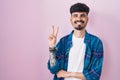 Young hispanic man with beard standing over pink background smiling with happy face winking at the camera doing victory sign Royalty Free Stock Photo