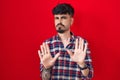 Young hispanic man with beard standing over red background moving away hands palms showing refusal and denial with afraid and Royalty Free Stock Photo